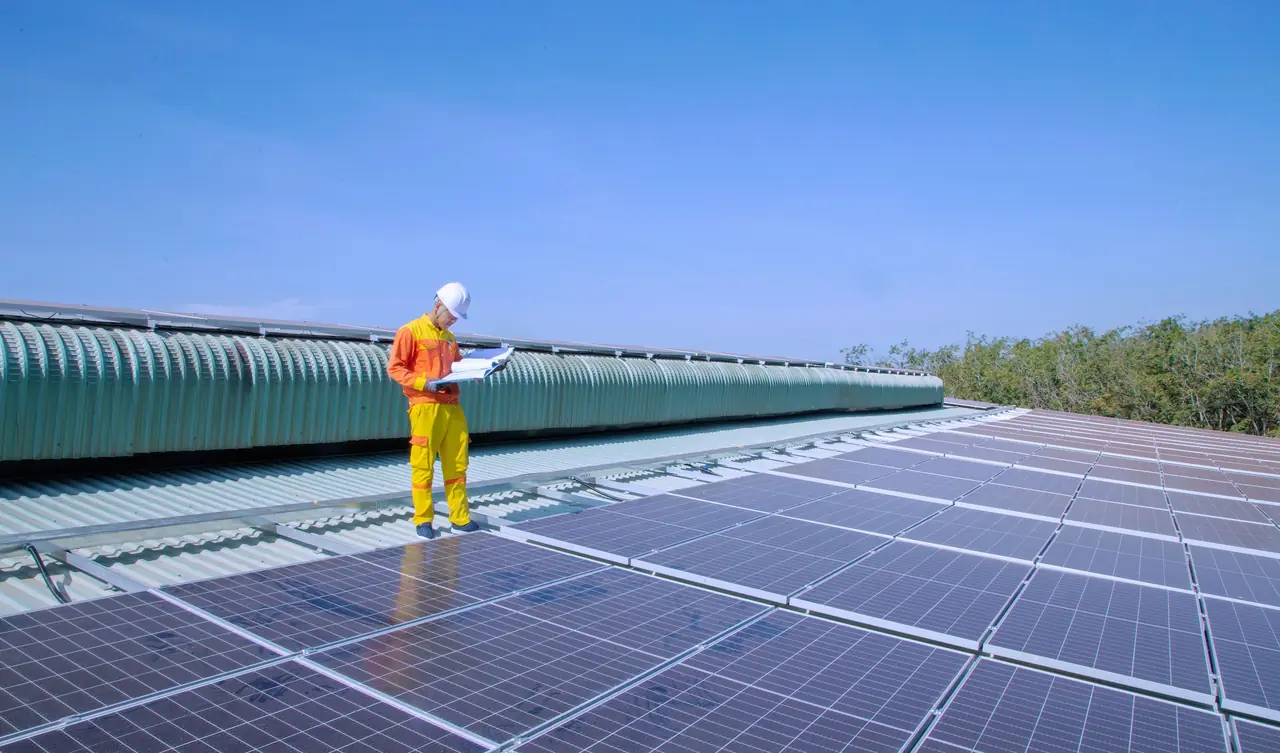 Técnico de pie sobre un techo cubierto de paneles solares revisando planos bajo un cielo despejado