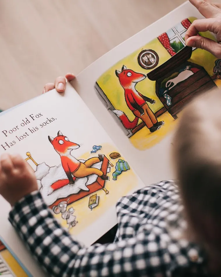 A caregiver and a child looking at an illustrated storybook together at a low table