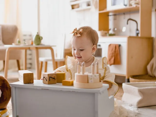 A baby playing with wooden number blocks and a toy cake on a low cabinet in a bright room