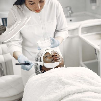 Esteticista con guantes aplicando una mascarilla a una clienta envuelta en toallas blancas